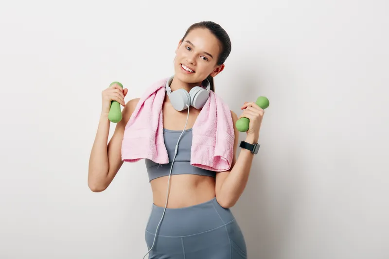 Fitness sport woman lifting dumbbells with a pink towel draped around shoulders, wearing workout outfit and headphones, smiling during a dynamic training session at home.