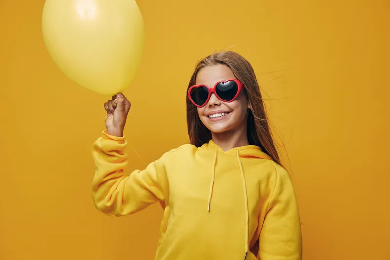 Girl smiling with heart shaped sunglasses and yellow balloon in hand, wearing yellow sweatshirt on yellow background for celebration and happiness concept.
