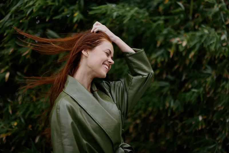 Портрет счастливой женщины с развевающимися рыжими волосами, улыбающейся с зубами, идущей по городу в парке на фоне зеленых листьев бамбука
