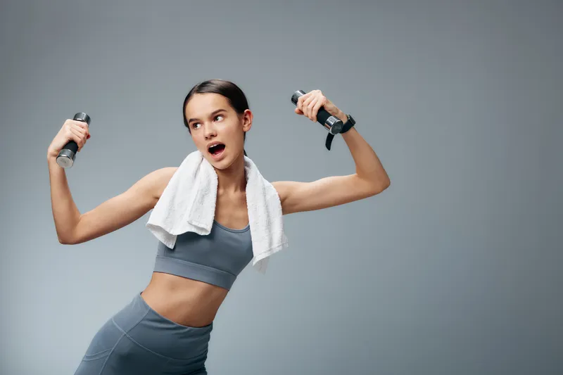 Female fitness enthusiast lifting dumbbells with a towel around her neck, delivering a dynamic workout pose, showcasing strength, energy, and athletic training in a studio setting.