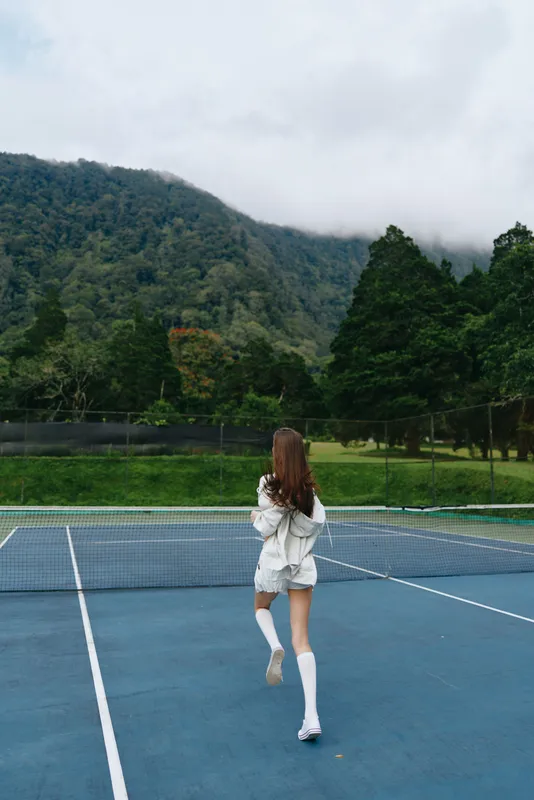 Young female tennis player in a gray hoodie and knee high socks, jogging on an outdoor court with a lush green background and mountains