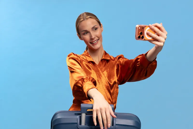 Smiling woman in orange blouse takes selfie with smartphone while holding suitcase handle on blue background. Concept of travel, vacation, and joyful moments captured on camera.