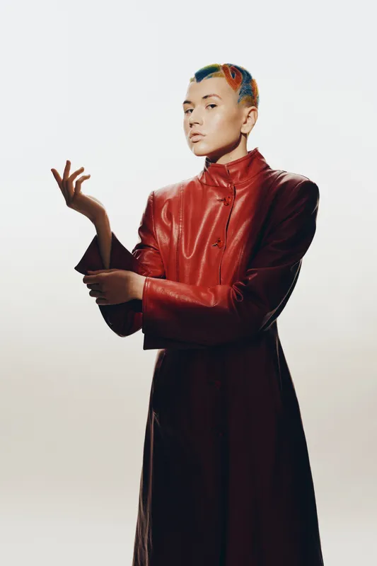 woman with colorful short hair wearing a red leather coat poses confidently against a light neutral background in a stylish contemporary fashion portrait.