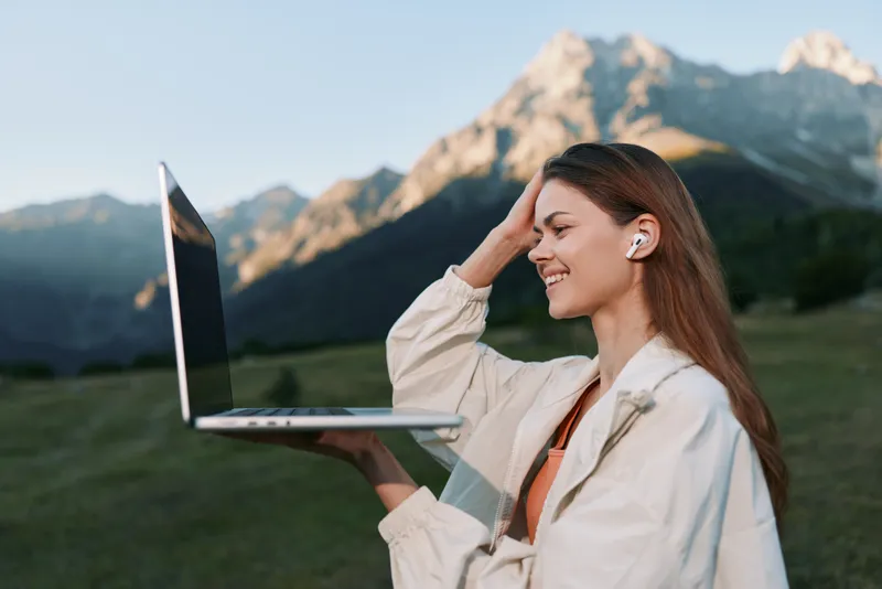 A cheerful female professional sits outdoors in a natural meadow, holding a laptop aloft as she smiles, enjoying a productive moment with scenic mountains in the background.