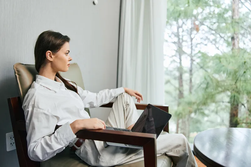 Молодая женщина в безупречном белом костюме удобно устроилась в современном кресле, работая за ноутбуком и наслаждаясь умиротворяющим видом на природу за окном