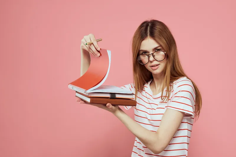 Молодая студентка с книгами на розовом фоне, в очках, крупный план лица