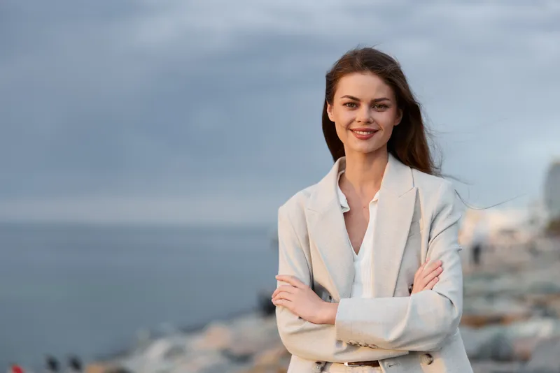 confident woman in light blazer standing outdoors with arms crossed by the sea on cloudy day, natural smile, casual business fashion style