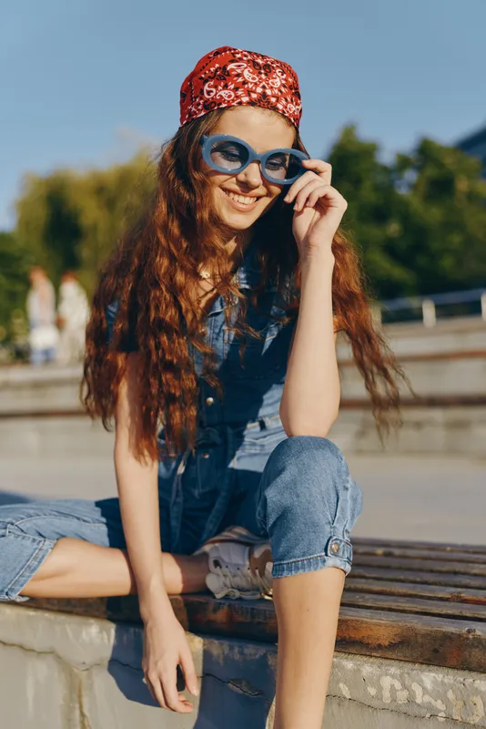 Lifestyle woman in modern boho-western denim street style wearing blue sunglasses and a red bandana, captured with warm film color tones in urban outdoor setting.