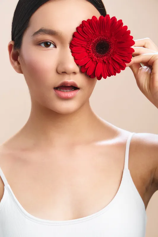 Asian woman holds a vivid red flower over her eye in a studio portrait, creating a bold, artistic mood with soft lighting and a clean minimalist background.