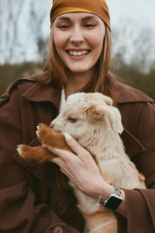 Smiling woman outdoors holds a fluffy goat kid, creating a lifestyle moment of affection, companionship, and nature, with a cozy atmosphere and soft light highlighting warmth and joy.