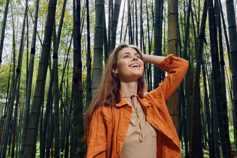 Woman in orange jacket stands in a bamboo forest smiling in a nature portrait, looking up with joy. Outdoors travel scene with relaxed lifestyle, sunlight and peaceful green woodland mood.