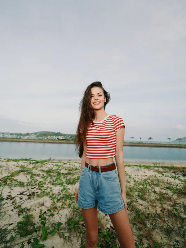Portrait of a smiling young woman standing on a sandy shoreline wearing a red and white striped crop top and denim shorts, with calm sea and distant horizon.