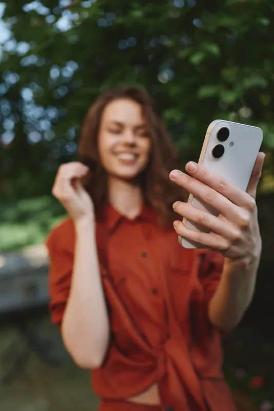Vibrant woman lifestyle moment outdoors, smiling while taking a selfie with smartphone, casual pose, sunlit greenery background, airy atmosphere, candid portrait, natural colors