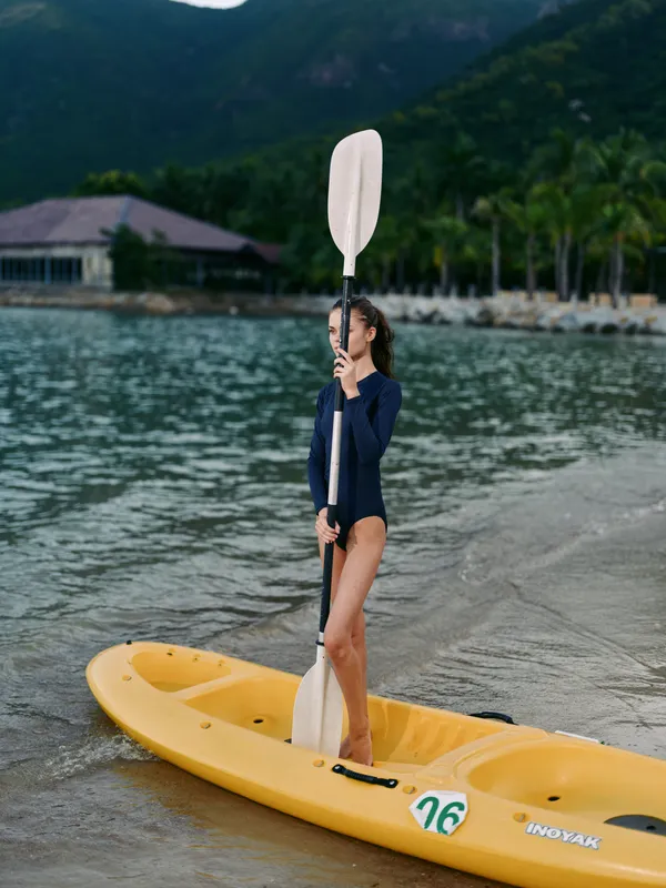 A person stands on a yellow kayak at the water's edge, gripping a paddle upright while calm waves lap the shore and distant mountains frame a serene outdoor water scene.