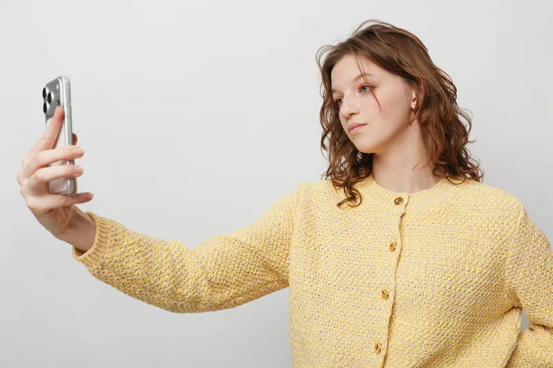 Young woman with curly hair wearing yellow knitted sweater takes selfie with smartphone on white background. Concept of casual style, social media, and modern communication.