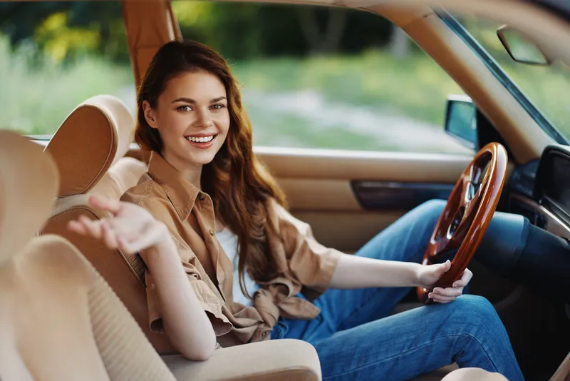 Young woman driving a car in a casual outfit, smiling in a natural outdoor setting, conveying joy and freedom.