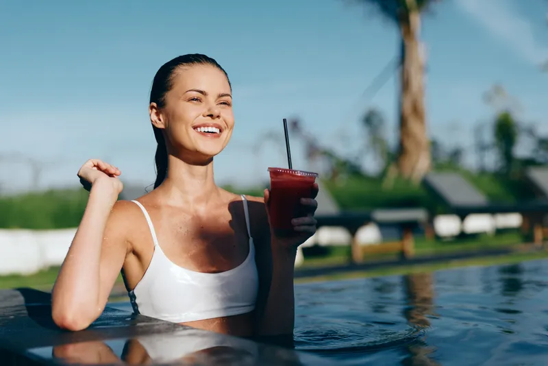 Smiling woman in a white swimsuit sits in a pool, holding a red smoothie with a straw. Bright outdoor setting conveys summer relaxation, wellness, and a carefree lifestyle