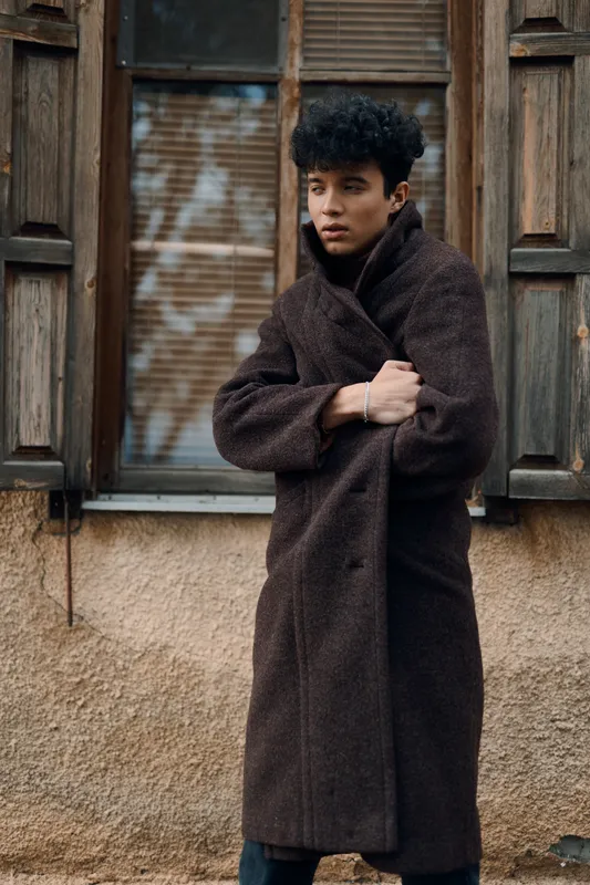 Модный портрет молодого человека в коричневом пальто, стоящего на фоне окна с руками на поясе и смотрящего в камеру