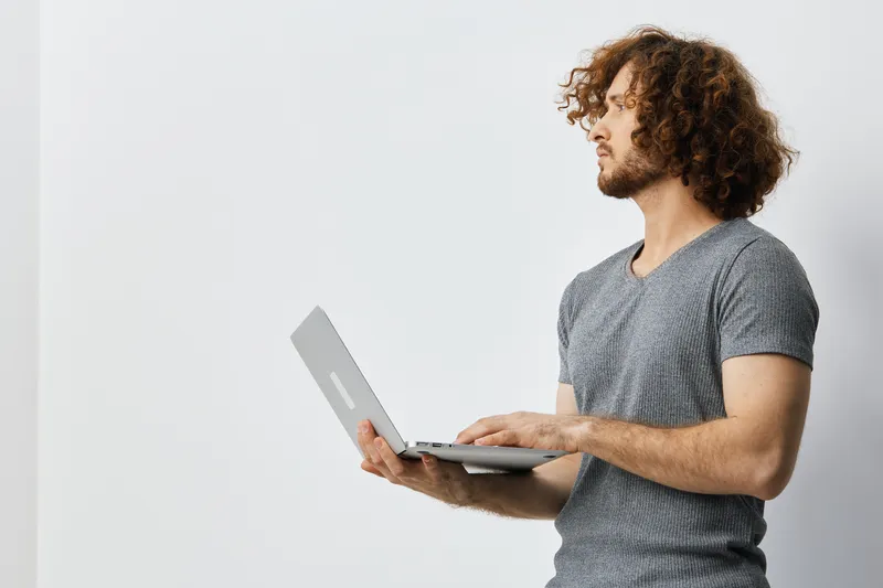 Thoughtful young man with curly hair working on a laptop, conveying focus and productivity in a minimalist workspace