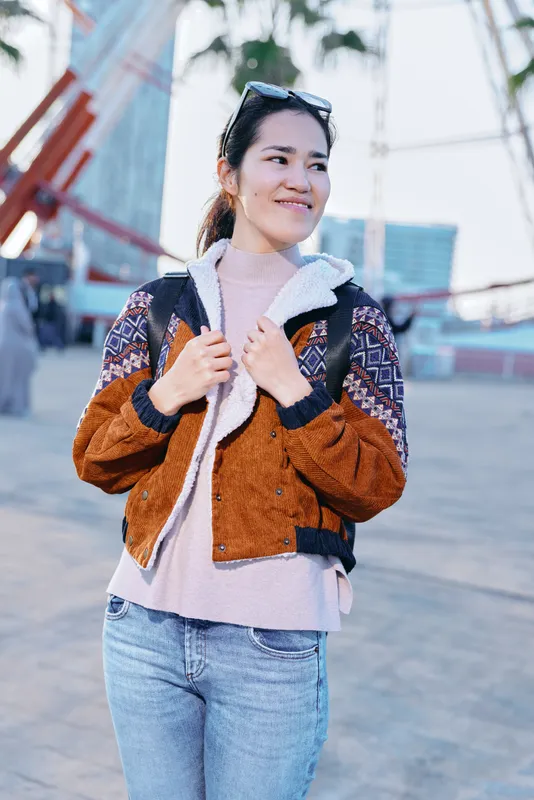 Woman in a warm brown fleece jacket over a pink sweater on an urban street, casual fashion, confident pose, sunny day, street style lifestyle photo