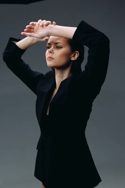 Serious woman in black blazer posing with arms raised in studio with dark background.