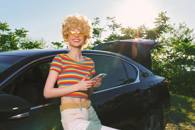 Cheerful woman outdoors, retro vibe, striped crop top, sunlit moment by a sleek car, relaxed pose, vibrant colors, natural background, playful mood, candid lifestyle scene