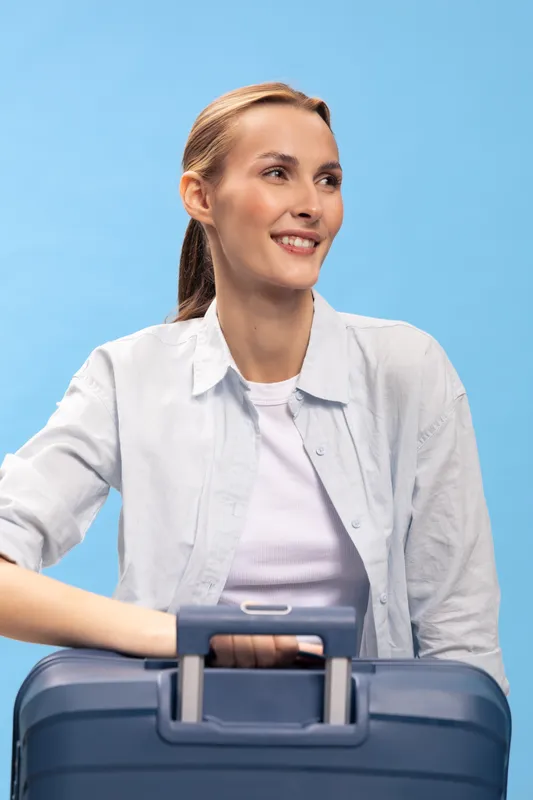 Young woman with light hair smiles holding blue suitcase handle. She wears white shirt and looks away on blue background. Concept of travel, vacation, and positive emotions.