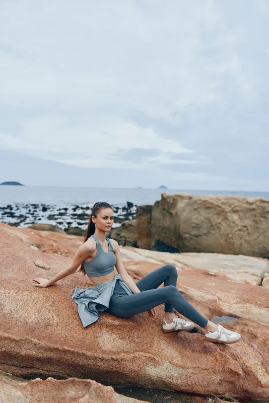 Fit young woman in gray activewear sitting on rocks by the sea, embodying a healthy lifestyle and tranquility against a scenic coastal background