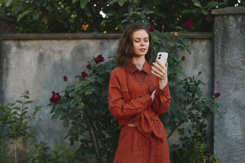 A young woman in a rust red belted dress stands in a garden, looking at her phone with a calm, focused expression, surrounded by blooming roses on a warm day
