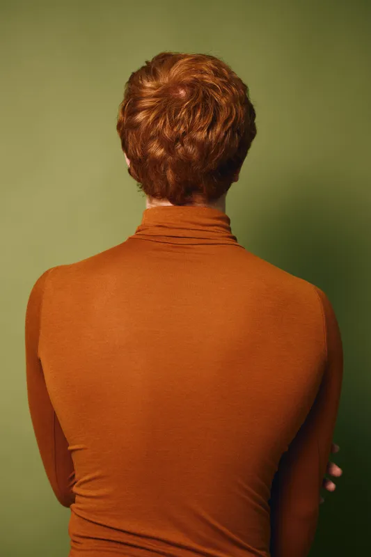 Man with red hair wearing brown turtleneck sweater standing with back turned against green background. Casual fashion, portrait, and hairstyle visible from behind with crossed arms.