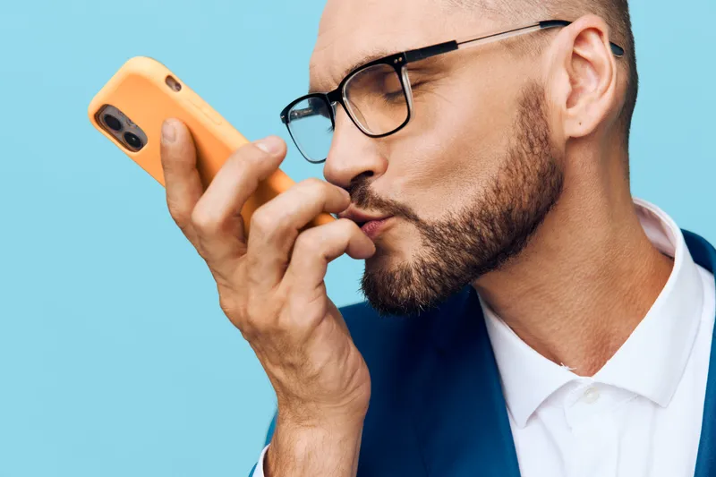 A professional man in business attire on a solid colored backdrop, expressing focused emotion while engaging with a mobile device.