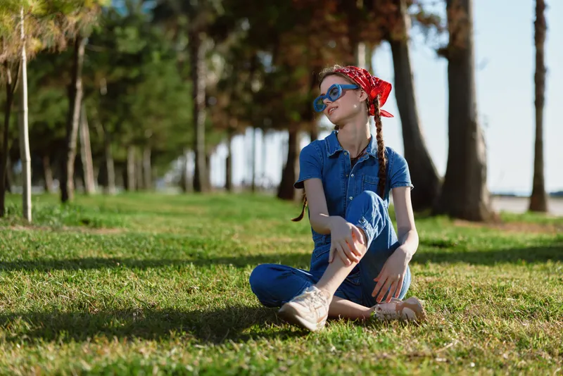 lifestyle woman in modern boho-western denim street style sitting on grass with braided hair, sunglasses, and red bandana, film color outdoors with trees background