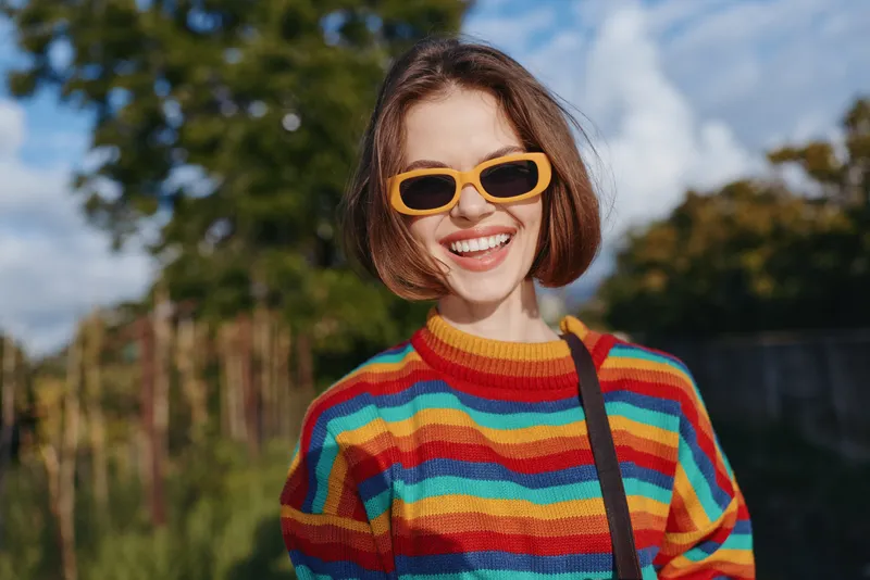 Young woman, smiling woman, rainbow sweater, yellow sunglasses, short bob, outdoor portrait – mid-20s model in casual outfit laughing in sunny park, colorful striped sweater, lifestyle travel vibe.