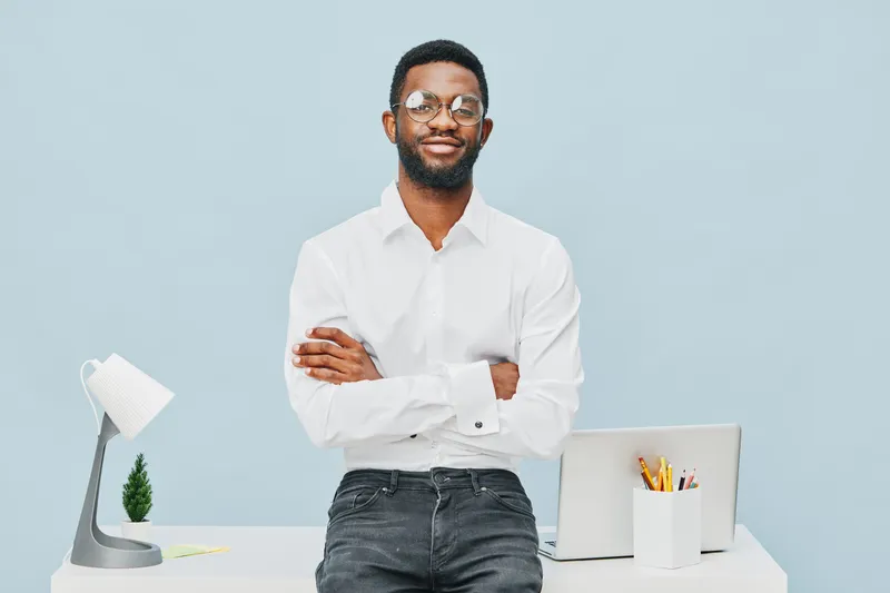 Confident young man with glasses standing at modern office desk crossed arms smiling, professional attire, minimalist workspace, light blue background, business concept