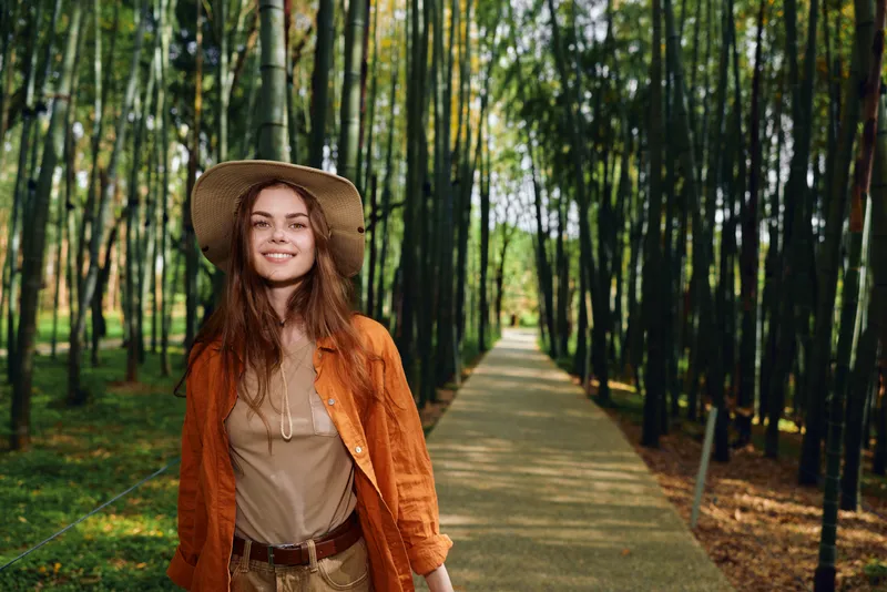 Woman in a bamboo forest on a path wearing a hat and smiling, portrait in nature. Casual travel scene walking along green trail with sunlight and relaxed joyful expression.