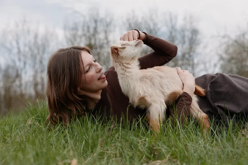 A woman lying on grass outdoors pets a playful goat kid, sharing a gentle moment of bond and trust. A lifestyle scene that highlights companionship between humans and animals in a natural setting.