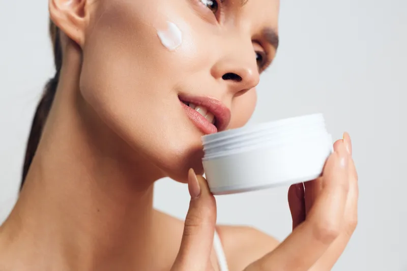 Young woman applies moisturizing cream on her cheek holds white jar close to face skincare beauty routine isolated on white background with natural soft light and smooth skin.