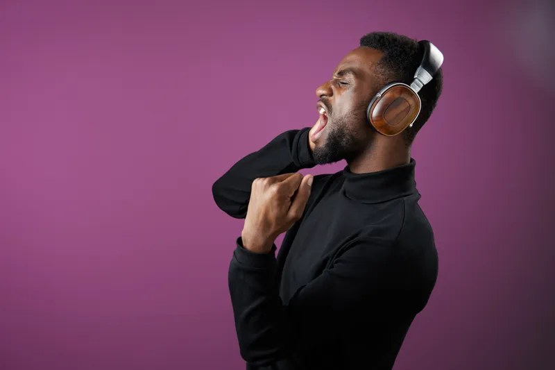 A joyful man in a black turtleneck sings passionately while wearing stylish headphones, expressing enthusiasm against a vibrant purple background