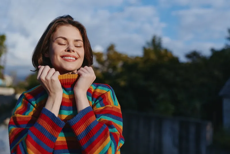 woman smiling young adult in a colorful striped sweater outdoors, eyes closed and holding collar, casual cozy autumn lifestyle portrait in a suburban backyard for travel and fashion.