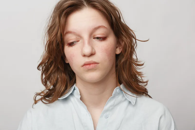 Sad young woman with wavy hair looks down wearing a light blue shirt on white background. Concept of loneliness, sadness, and emotional struggle in youth.