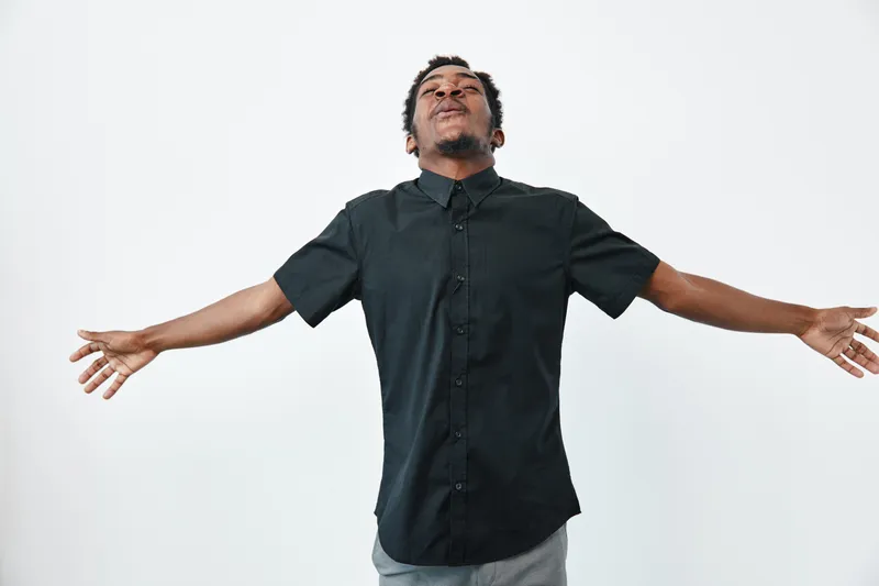 African man with dark skin wearing black shirt standing with arms wide open and eyes closed on white background. Male adult expressing freedom and relaxation in casual clothing.