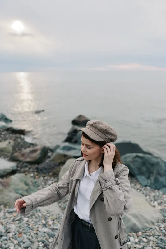 Young woman in beige coat and patterned cap stands on rocky shore near calm sea under cloudy sky. Concept of autumn fashion, nature, and peaceful outdoor lifestyle.