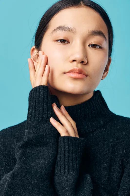 Portrait of young woman with smooth skin and dark hair wearing black turtleneck sweater posing against blue background with calm expression and hand touching face for beauty concept.