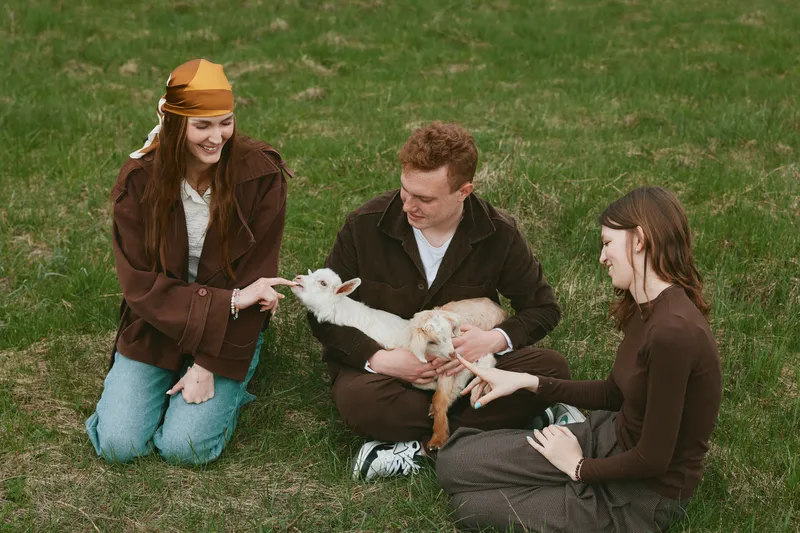 Three adults sit on green grass outdoors, sharing a tender moment with a small white goat kid, smiling and gently petting it, highlighting lifestyle, animal bonding, and wholesome countryside vibes.