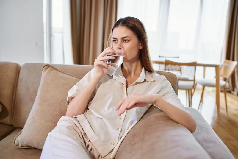 Woman drinking water from a glass on a sofa at home, relaxed casual pose with natural light, healthy hydration and comfort in a bright livingroom interior.