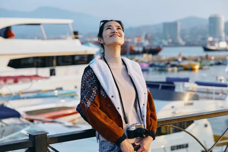 Woman in a cozy jacket stands by the marina, looking up with a content smile as boats drift and city skyline glows behind, a relaxed seaside lifestyle moment in outdoor fashion.