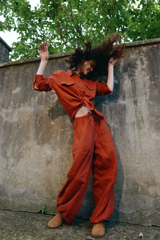 Energetic woman dances outdoors wearing a red jumpsuit against a weathered wall, capturing movement and casual street style in a bright daytime scene