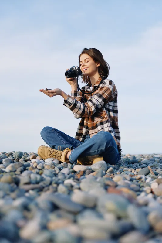 Woman photographer sitting on a pebble beach with a camera raised, capturing coastal scenery in natural light, wearing a plaid shirt and jeans for a relaxed outdoor moment