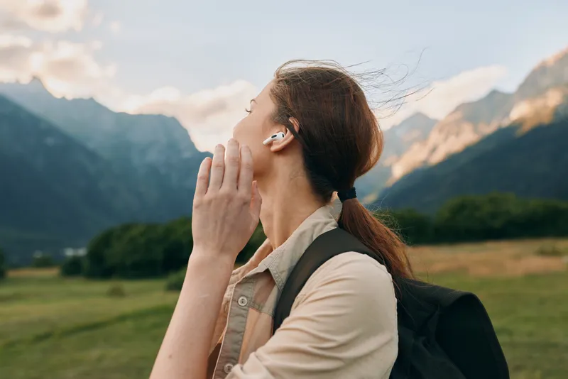 Young woman with long brown hair enjoys nature while listening to music with wireless earbuds during a hike in the mountains at sunset.