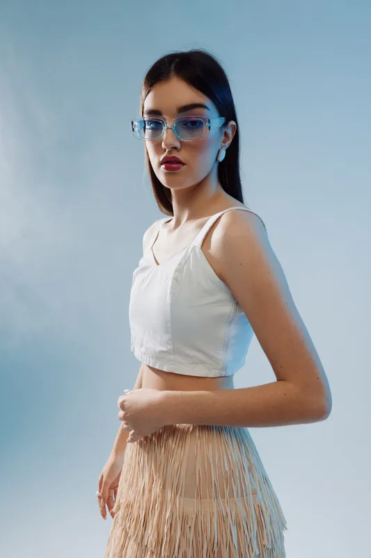 Young woman with long dark hair wearing glasses and a white top poses against a blue background. Concept of beauty, fashion, and modern style in studio lighting.